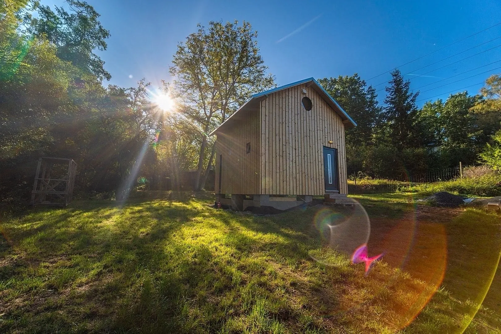 Dřevěný domek se svislou fasádou a sedlovou střechou na zahradě, v protisvětle